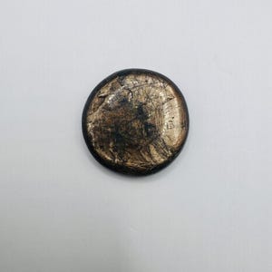 May include: A circular button with a dark, metallic bronze finish. The surface features a distressed, textured appearance with visible black streaks and scratches across the center, while the outer edge is smooth and dark.