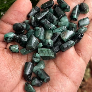 May include: A handful of raw emerald gemstones in various shapes and sizes. The emeralds are a deep green color, with some showing dark inclusions. The gemstones are rough-cut and appear to be natural, with a slightly polished surface.