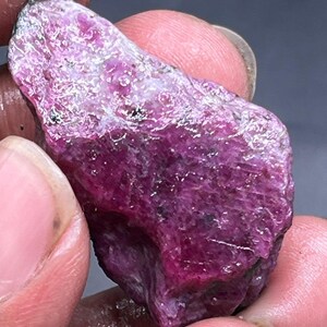 May include: A rough, uncut ruby gemstone with a deep magenta hue. The surface of the stone has a textured appearance with areas of lighter pink and white. The ruby is held between fingers, showcasing its natural, raw form. This gemstone is ideal for jewelry making or collecting.