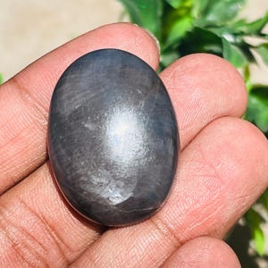May include: A polished, oval-shaped gemstone with a dark gray color and a subtle sheen. The stone is held in a hand, with the natural light reflecting off its surface. The gemstone's smooth, rounded form is clearly visible.