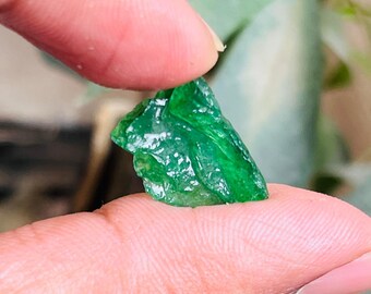 May include: A small, rough, green gemstone with a translucent appearance. The gemstone is held in a hand.