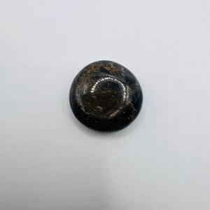 May include: A smooth, circular, polished stone with a dark black base color featuring internal golden-brown fibrous inclusions that create a reflective, chatoyant effect across the surface of the rounded, flattened gemstone.