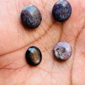 May include: Four cabochon gemstones, two are round and two are oval. The round gemstones are purple with black inclusions. The oval gemstones are black with gold inclusions.