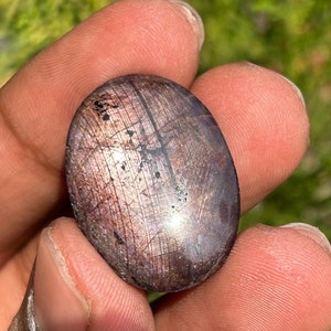 May include: A close-up of a purple and silver oval-shaped gemstone with black inclusions. The gemstone is smooth and polished.