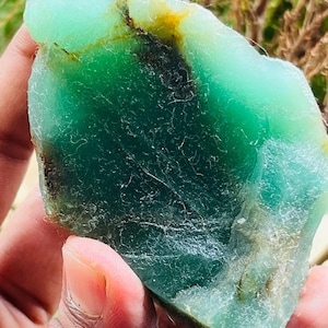 Puede incluir: Una gema de crisoprasa natural en bruto, con un tono verde vibrante, acentuado por inclusiones verdes más oscuras y marrones. La piedra tiene una superficie rugosa y texturizada. Se muestra en una mano, resaltando su belleza natural.