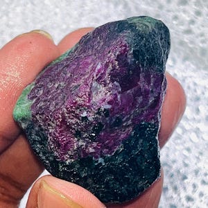 Puede incluir: Una gema en bruto con un centro púrpura intenso, que se transforma en bordes verde oscuro y negro. La piedra tiene una superficie texturizada y se sostiene en una mano, mostrando su belleza natural y en bruto. Un espécimen mineral único.