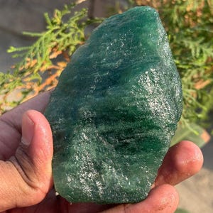 May include: A raw, unpolished piece of green aventurine gemstone. The natural stone has a textured surface and varying shades of green. The gemstone is held in a hand, with a blurred background of green foliage.