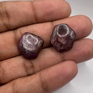 May include: Two raw, hexagonal-shaped ruby gemstones with a deep reddish-purple hue. The stones feature a natural, unpolished texture with visible surface striations and internal inclusions, showcasing a semi-opaque appearance and earthy finish.