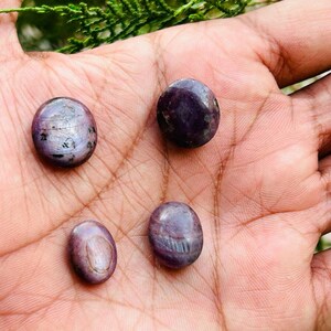 May include: Four purple and grey gemstone cabochons, likely ruby, in various shapes and sizes, displayed on a hand.