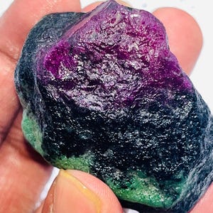 Puede incluir: Una gema en bruto sin cortar con un llamativo degradado de color. La parte superior es de un púrpura vibrante, que pasa a un negro profundo y termina con un toque de verde en la base. La textura rugosa de la piedra es visible.