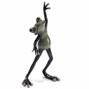 May include: A green and black ceramic frog figurine with one leg raised in the air. The frog has large, black eyes with gold accents and a wide, toothy grin.