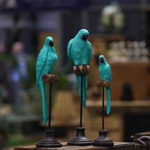 May include: Three teal blue parrot figurines on black stands. The parrots are perched on small branches. The figurines are made of a smooth, painted material.