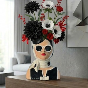 May include: A decorative vase in the shape of a woman's head, wearing sunglasses and a polka dot scarf. The vase holds black dahlias, white anemones, red roses, and red berries. The vase is dressed in a black and white outfit.