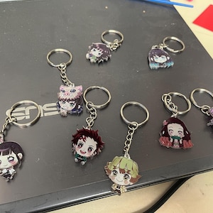 May include: Collection of anime character keychains. Each keychain features a colorful, detailed illustration of a character's face. The keychains have silver-colored metal rings and chains. The characters have various hair colors and expressions.