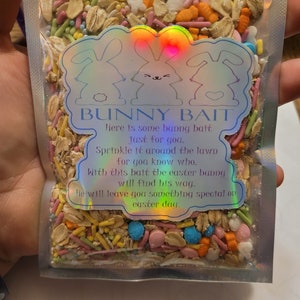 Op de afbeelding: Een doorzichtige plastic zak gevuld met kleurrijke hagelslag, waaronder roze, oranje, groen en blauw. De zak heeft een holografisch label met konijnenillustraties en de tekst "BUNNY BAIT". De tekst op het label luidt "Hier is wat konijnenaas voor jou."