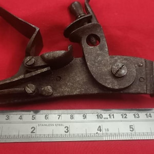 May include: A close-up of a rusty metal gun lock mechanism with a ruler for scale. The lock is approximately 15 cm long and has a variety of moving parts.