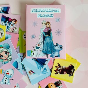 Puede incluir: Una caja rosa con el texto "Memorarama Frozen" en la parte delantera. La caja presenta una imagen de Elsa, Anna, Olaf y una salamandra azul. La caja está rodeada de pequeñas tarjetas cuadradas con imágenes de personajes de la película Frozen.