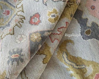 May include: Close-up of a rolled-up rug with a detailed floral pattern. The rug features a light gray background with accents of gold, pink, and dark gray. The texture is visible, showcasing the woven construction. The text "RigaRugs" is visible in the bottom right corner.