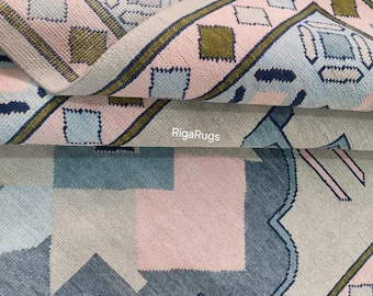 May include: Close-up of a blue, pink, and beige patterned rug with a geometric design. The rug has a fringe border and the text "Riga Rugs" is visible in the image.