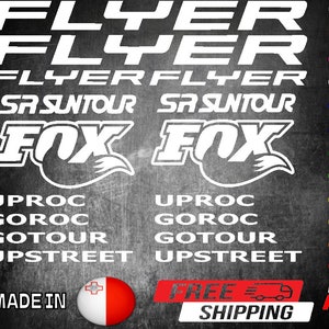 Pode incluir: Adesivos brancos com as palavras "FLYER", "SR SUNTOUR" e "FOX" em uma fonte em negrito sem serifa. Os adesivos também incluem as palavras "UPROC", "GOROC", "GOTOUR" e "UPSTREET". Os adesivos são exibidos em um fundo cinza escuro.