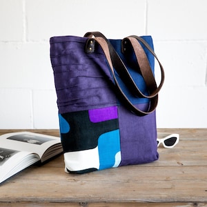 Può includere: Una borsa tote in patchwork viola e blu con manici in pelle. La borsa presenta un motivo geometrico in tonalità di blu, bianco e viola.