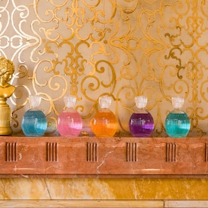 May include: Six round glass bottles with colorful liquids on a marble shelf in front of a gold patterned wall. The bottles have clear glass stoppers. A gold statue of a woman's bust is on the shelf to the left of the bottles.
