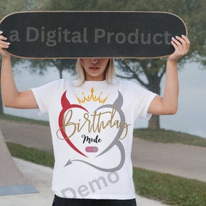 Puede incluir: Camiseta blanca con un diseño de corazón gris y rojo. El corazón tiene cuernos de diablo y una corona dorada en la parte superior. El texto "Birthday Mode" está dentro del corazón. La palabra "Demo" está impresa en la parte inferior de la camiseta.