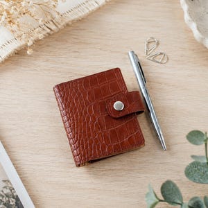 May include: A small, rectangular brown wallet featuring a textured crocodile skin pattern and a secure snap-button closure. The wallet is positioned next to a sleek silver ballpoint pen, creating a compact set for daily organization and note-taking.