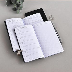 Pode incluir: Cadernos pretos abertos com papel quadriculado, um mostrando um layout de calendário semanal de fevereiro de 2024. Uma caneta prateada e clipes de papel estão por perto. Os cadernos estão em uma superfície cinza.