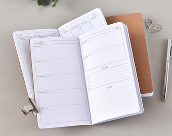May include: An open planner with brown covers featuring a weekly layout for January 2026, including sections for To Do lists and notes. A silver metal clip is attached to the left page, and a silver pen lies to the right of the planner.