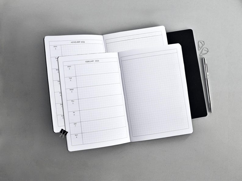 May include: Two open planners with white pages featuring a grid layout and weekly calendar sections labeled February 2024 and November 2024. A black notebook cover, a silver pen, and two small silver paper clips are positioned nearby.