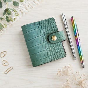 Può includere: Un piccolo portafoglio verde con una trama testurizzata a pelle di coccodrillo, dotato di una chiusura a bottone a pressione dorato. Accanto al portafoglio ci sono due penne, una argentata e una iridescente arcobaleno, e tre graffette dorate.