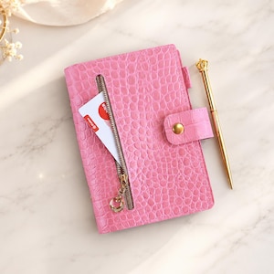 May include: A bright pink notebook with a textured crocodile skin pattern, featuring a vertical zippered pocket on the front containing a white card, a snap-button closure, and a gold-toned pen with a crown-shaped topper resting beside it.