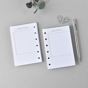 May include: Two white planner pages with lined sections and time slots, dated January 22 and 23, lie on a gray surface. A silver pen and two silver paper clips are also present. The pages have holes for binding.