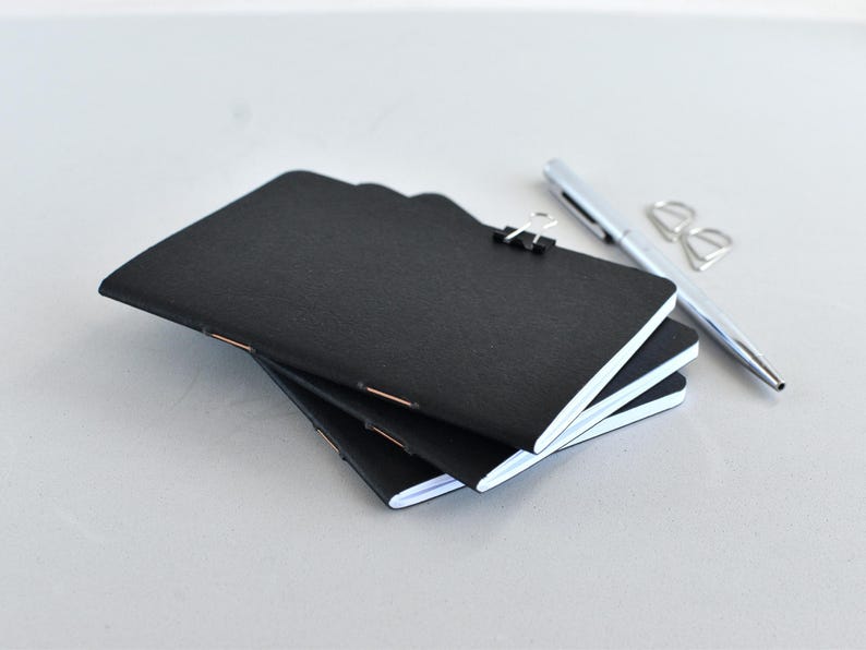 May include: A stack of three small black notebooks with white pages, bound with copper-colored staples. A black metal binder clip secures the top notebook. A silver ballpoint pen and a small silver metal paper clip lie beside the notebooks.