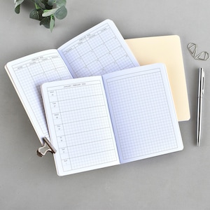Könnte beinhalten: Geöffnete Notizbücher mit kariertem Papier und Kalenderseiten, zusammengehalten von einer silbernen Klammer. Ein silberner Stift und Büroklammern liegen neben den Notizbüchern. Die Notizbücher sind geöffnet, um die Kalender- und Wochenplanseiten zu zeigen.