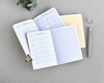 Puede incluir: Cuadernos abiertos con papel cuadriculado y páginas de calendario, sujetos con un clip plateado. Un bolígrafo plateado y clips están junto a los cuadernos. Los cuadernos están abiertos para mostrar las páginas del calendario y del planificador semanal.