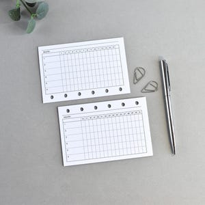 Può includere: Due fogli bianchi per planner con layout a griglia e giorni numerati, insieme a una penna d'argento e graffette. I fogli hanno fori preforati per la rilegatura. Su una superficie grigia.