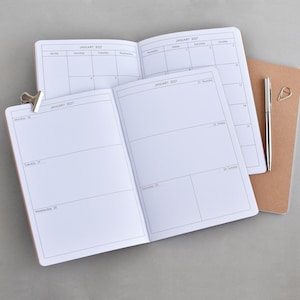 Op de afbeelding: Open notitieboekjes met lege pagina's en kalenderindelingen. Een zilveren pen en een gouden paperclip liggen op een bruin notitieboek. Een zilveren clip houdt de pagina's bij elkaar. De notitieboekjes zijn open voor de maand januari 2027.