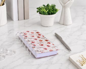 May include: A small disc-bound notebook with a white cover featuring a repeating pattern of red and pink circles and moon phase icons. The notebook has clear plastic binding discs and is accompanied by a silver metal ballpoint pen.