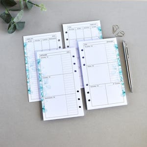 Könnte beinhalten: Drei weiße Planer-Einlagen mit blauem Aquarell-Blumenrand, die schwarzen Text für Wochentage und Daten enthalten. Die Seiten haben links Lochungen für einen Ordner. Ein silberner Metallkugelschreiber und zwei Büroklammern sind ebenfalls abgebildet.