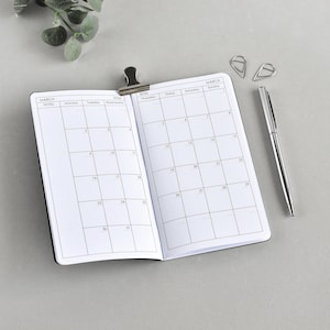 Könnte beinhalten: Offenes Notizbuch mit einem Kalender für März 2024, gehalten von einer schwarzen Klammer. Ein silberner Stift und zwei silberne Büroklammern liegen daneben. Der Hintergrund ist eine hellgraue Oberfläche.