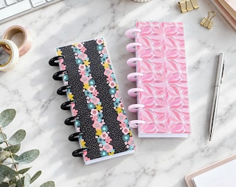 May include: Two slim disc-bound notebooks. One notebook features a black cover with a vertical pattern of pink, yellow, and teal flowers and white dots. The second notebook has a pink cover with a repeating geometric floral pattern.