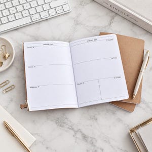 Könnte beinhalten: Ein offener Planer mit weißen Seiten, der ein wöchentliches Kalenderlayout für Januar 2027 mit horizontalen Linien für tägliche Einträge zeigt. Der Planer hat einen braunen Einband und wird von einem weißen Stift und einem braunen Notizbuch begleitet.