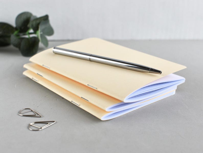 May include: A stack of three cream-colored notebooks with staple-bound spines, topped with a silver metallic ballpoint pen. Two silver triangular paper clips are positioned to the side of the notebooks.