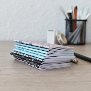 Könnte beinhalten: Ein Stapel aus sechs schmalen Notizheften mit verschiedenen gemusterten Einbänden, darunter hellrosa, hellblaue, florale und schwarze Designs. Die Hefte sind mit Klammern geheftet und haben weiße Papierseiten. Zwei goldene herzförmige Büroklammern.