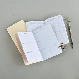 Puede incluir: Dos agendas abiertas con páginas blancas que muestran calendarios y horarios semanales impresos en negro para febrero de 2027. Un bolígrafo de metal plateado y un clip de carpeta plateado se colocan junto a las agendas de cubiertas color crema.