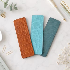 May include: Three rectangular bookmarks featuring a textured crocodile skin pattern. The bookmarks are colored in shades of burnt orange, bright turquoise, and muted teal, each with rounded corners and a flat, slim profile for page marking.