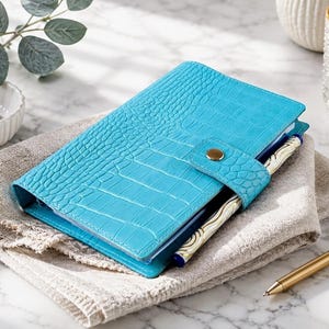 May include: A bright turquoise planner with a textured crocodile skin pattern, featuring a gold-toned snap button closure. The planner includes a pen holder containing a white and gold patterned pen, and it is resting on a folded beige fabric cloth.