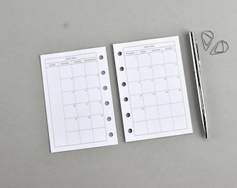 May include: Two white planner inserts for April 2024 with a grid layout, a silver pen, and two silver paper clips on a gray surface. The inserts have pre-punched holes for binding.